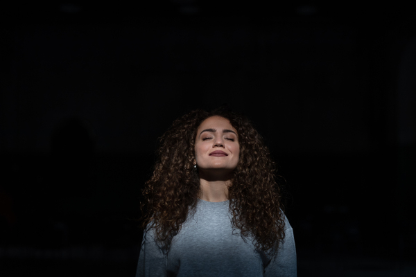 A portrait of young woman standing indoors against black bacground, eyes closed.