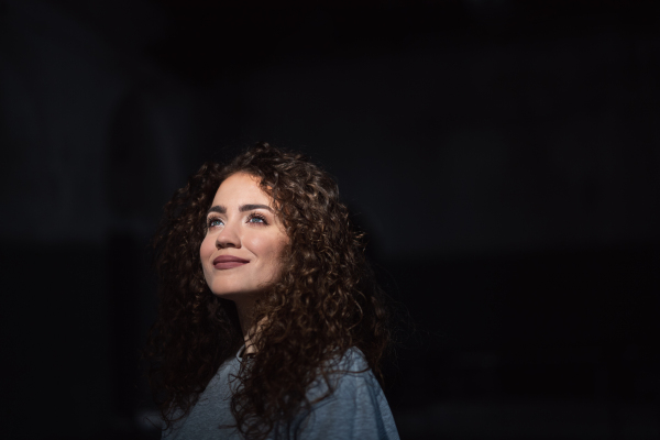 A portrait of young woman standing indoors against black bacground, looking up.