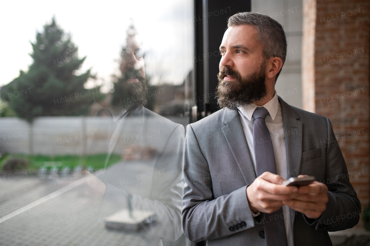 A portrait of mature businessman standing indoors, using smartphone.