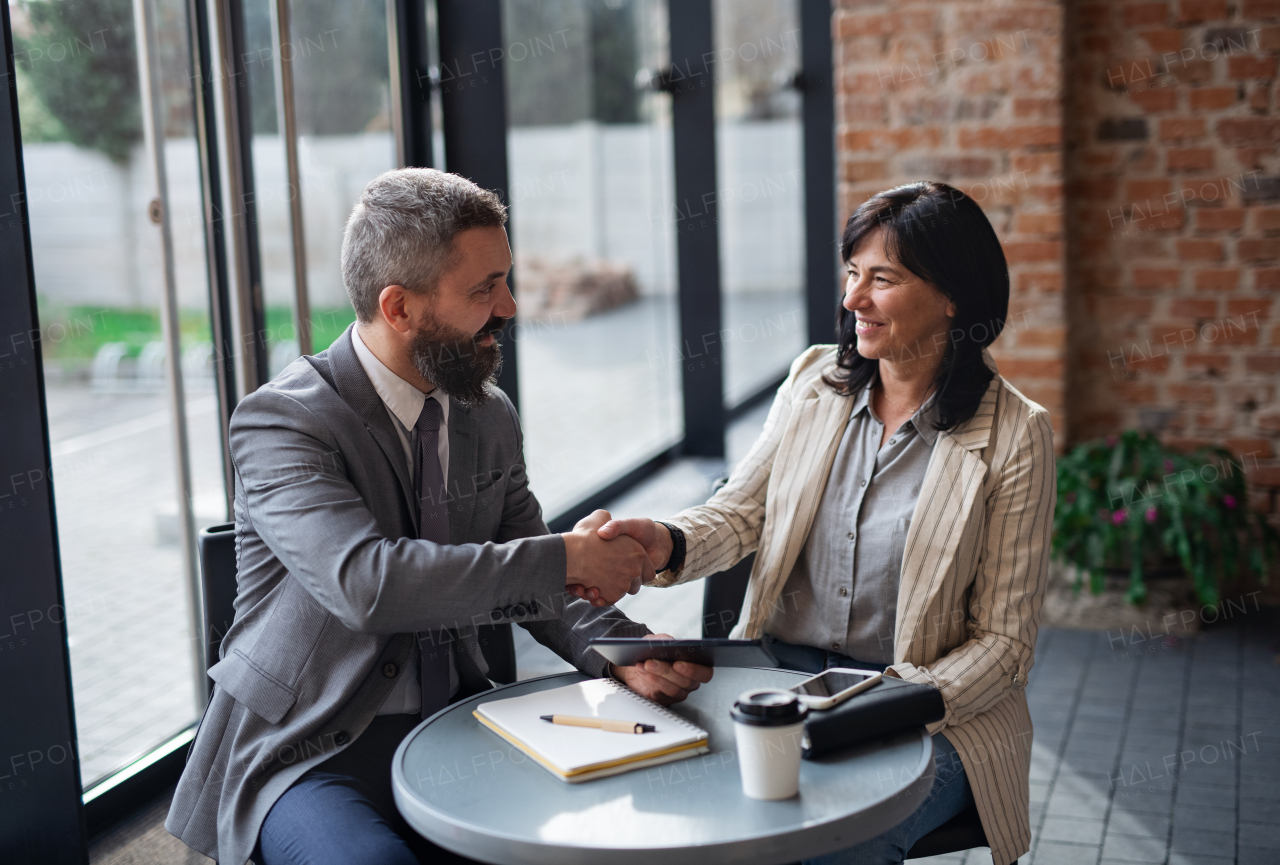 A portrait of businesspeople sitting at the table and working indoors, shaking hands.