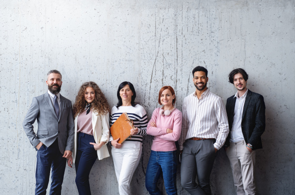 Front view portrait of group of entrepreneurs standing against concrete wall indoors in office, looking at camera.