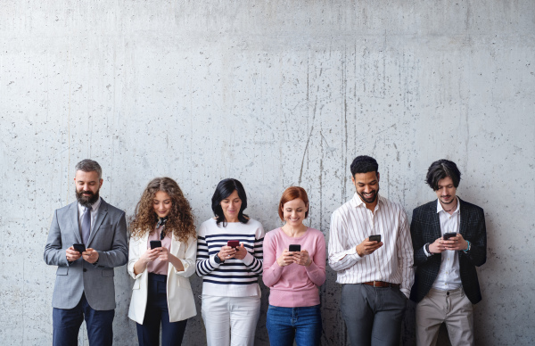 Front view portrait of group of entrepreneurs standing against concrete wall indoors in office, using smartphone.