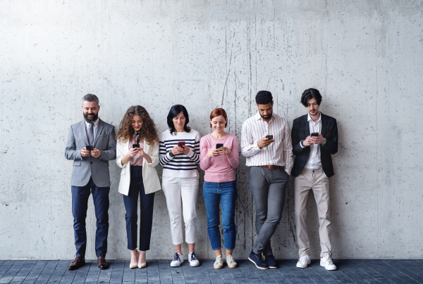 Front view portrait of group of entrepreneurs standing against concrete wall indoors in office, using smartphone.