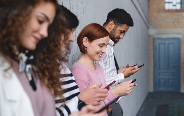 Side view of group of entrepreneurs standing against concrete wall indoors in office, using smartphone.