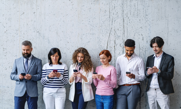 Front view portrait of group of entrepreneurs standing against concrete wall indoors in office, using smartphone.