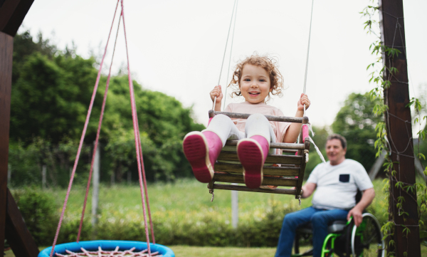 Happy small girl with senior grandfather in wheelchair playing in backyard garden, swinging.