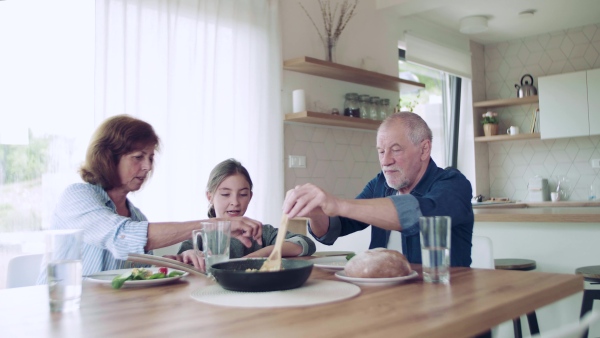 A happy small girl with senior grandparents indoors sitting at the table, eating.