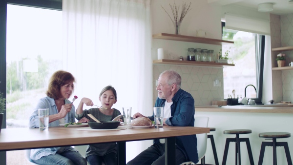 A happy small girl with senior grandparents indoors sitting at the table, eating.