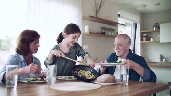 A happy small girl with senior grandparents indoors sitting at the table, eating. Slow motion.