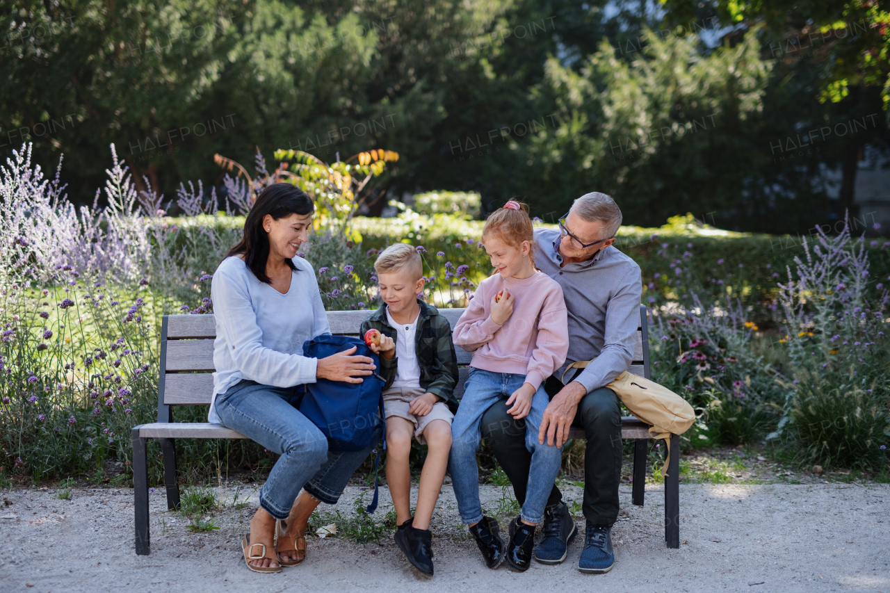 A senior couple with grandchildren sitting on bench, eating snack and talking outdoors in park.