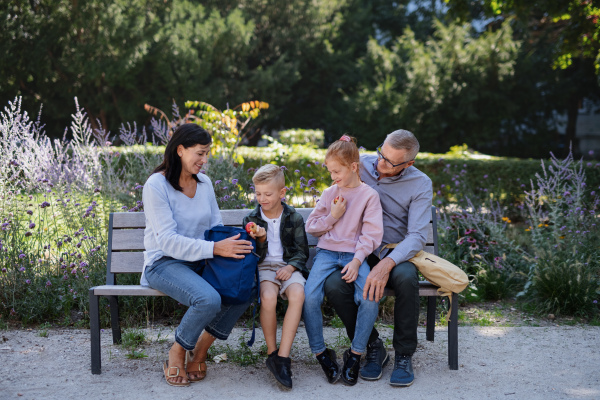 A senior couple with grandchildren sitting on bench, eating snack and talking outdoors in park.