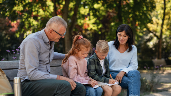 A senior couple with grandchildren sitting on bench and doing homewrok outdoors in park.