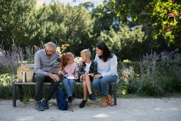 A senior couple with grandchildren sitting on bench and doing homewrok outdoors in park.