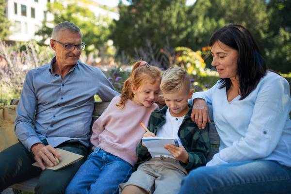 A senior couple with grandchildren sitting on bench and doing homewrok outdoors in park.