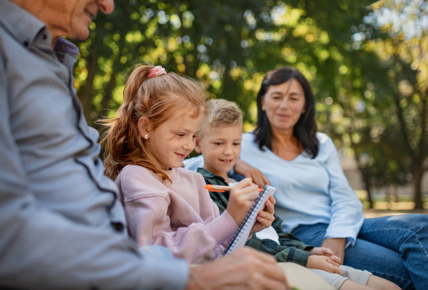 A senior couple with grandchildren sitting on bench and doing homewrok outdoors in park.