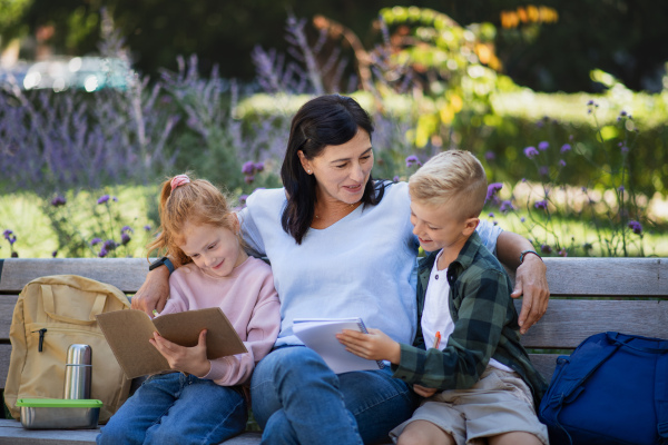 A happy senior woman with grandchildren sitting on bench and helping with homework outdoors in park.