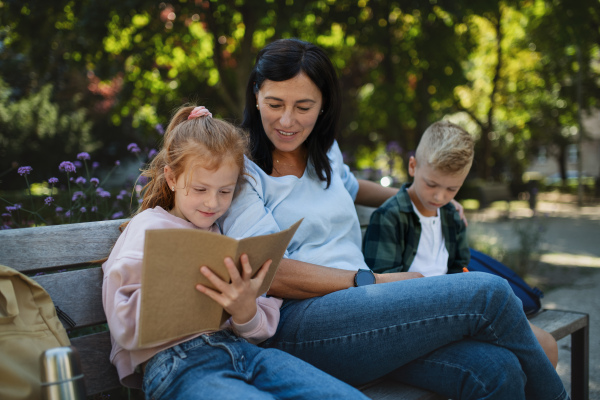 A happy senior woman with grandchildren sitting on bench and helping with homework outdoors in park.