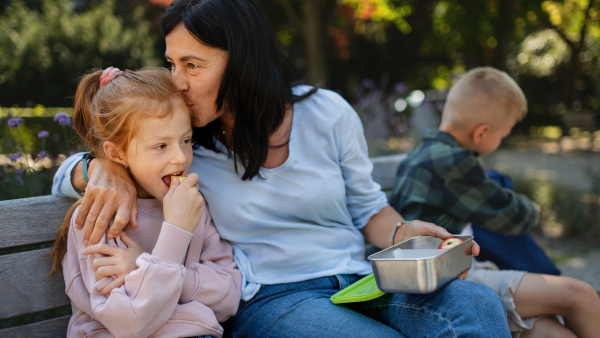 A happy senior woman sitting on bench and giving kiss and snack to her granddaughter outdoors in park.