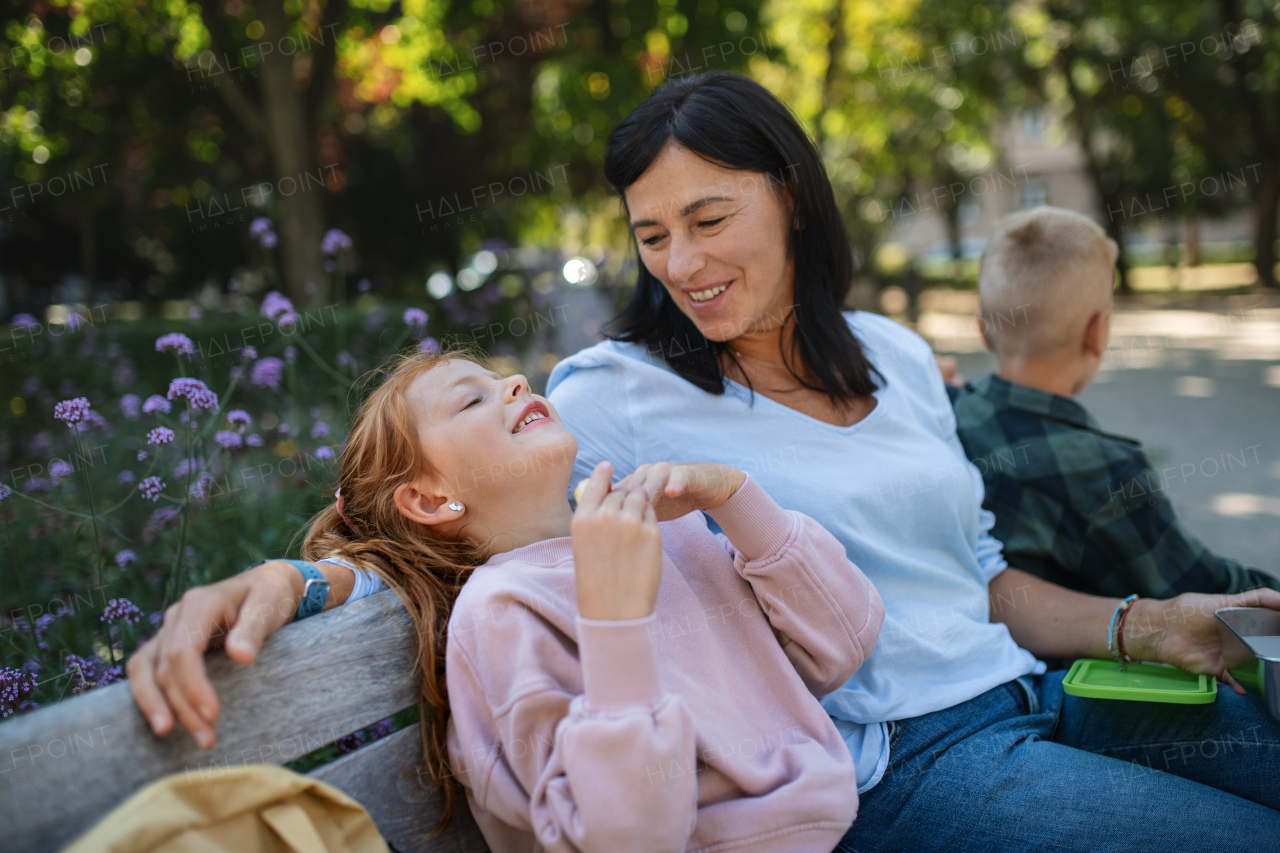 A happy senior woman with grandchildren sitting on bench and having snack outdoors in park.