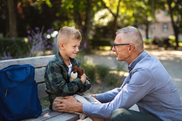 A senior man squatting and talking to grandson outdoors on bench in park.