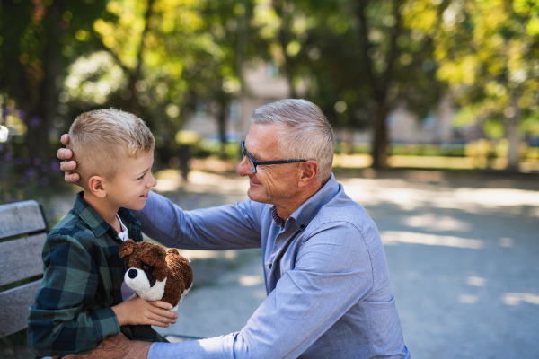 A senior man squatting and talking to grandson outdoors on bench in park.