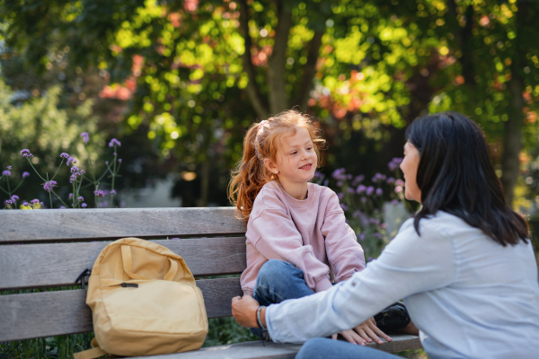 A senior woman squatting and talking to granddaughter outdoors on bench in park.