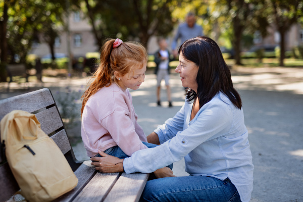 A senior woman squatting and talking to granddaughter outdoors on bench in park.