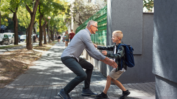 A happy schoolboy running to his grandfather waiting for him after school outdoors in street.