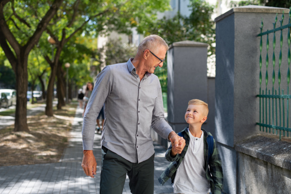 A happy grandfather taking his grandson home from school, walking outdoors in street.