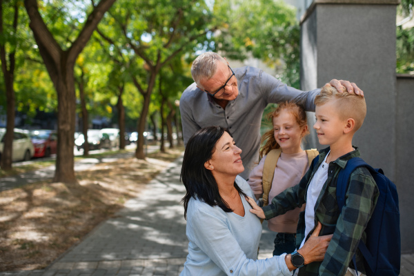 Happy grandparents taking grandchildren home from school, waiting in front of a school outdoors in street.