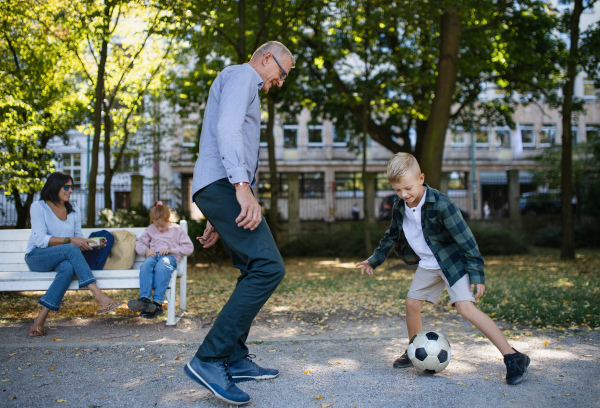 A little boy playing football with granddad, his sister and grandmom sitting on bench outdoors in park