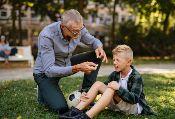 A little boy with injured leg crying, his grandfather is giving him plaster outdoors in park.