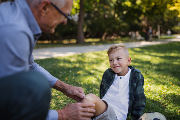 A little boy with injured leg getting plaster from grandfather outdoors in park.