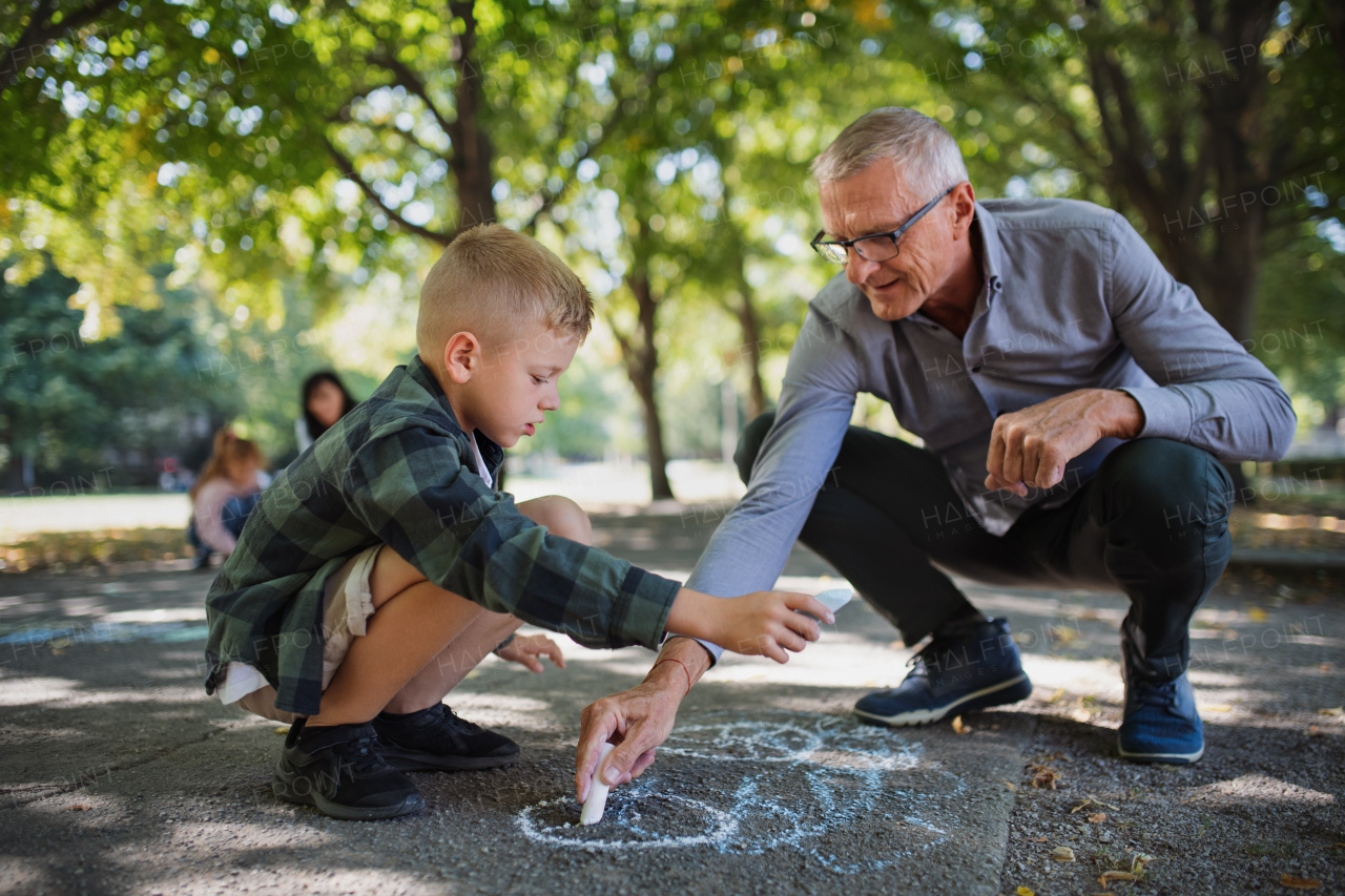 A grandfather with grandson drawing with chalks on pavement outdoors in park.