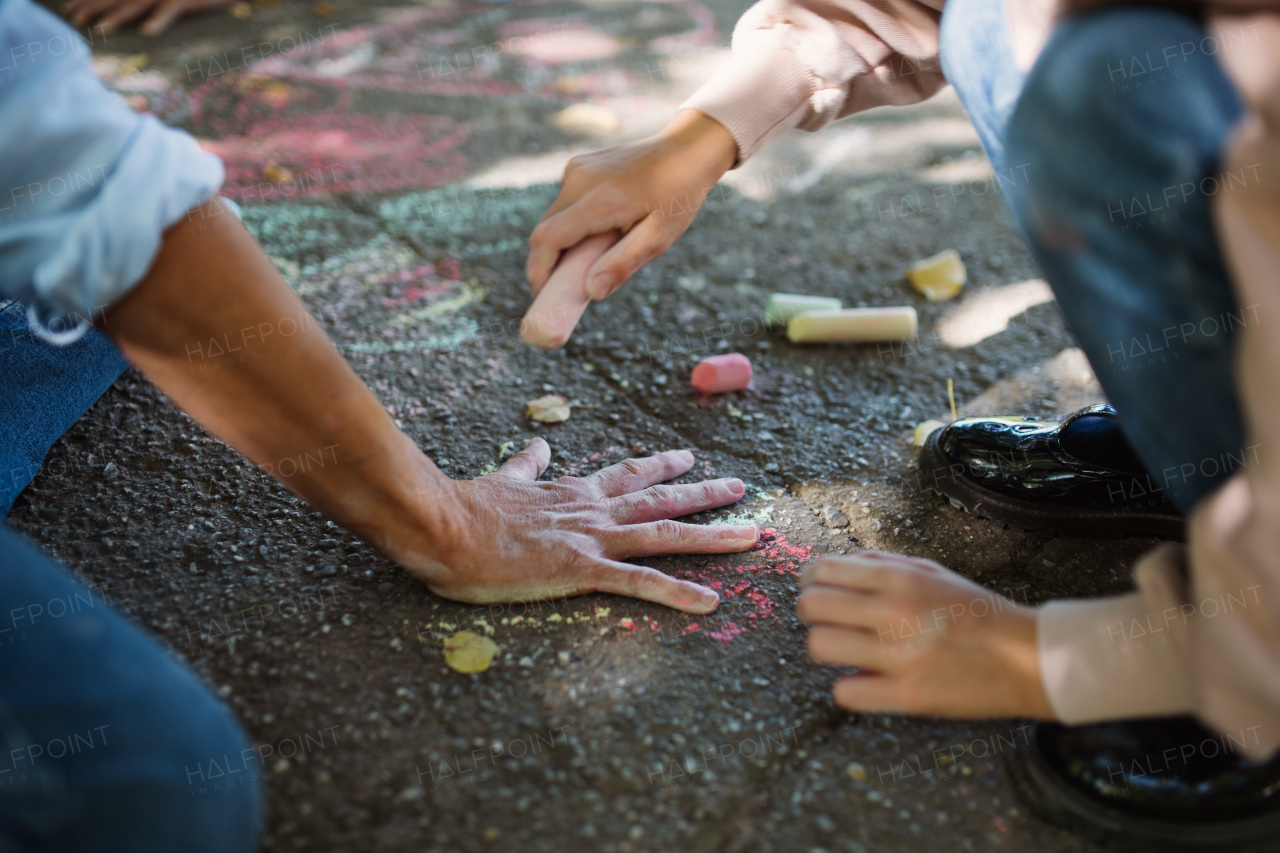 An unrecognizable little girl drawing grandmother's hand with chalk on pavement outdoors.