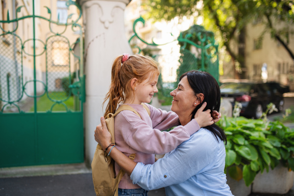 A happy schoolgirl hugging his grandmother waiting for her after school outdoors in street.