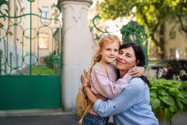 A happy schoolgirl hugging his grandmother waiting for her after school outdoors in street.