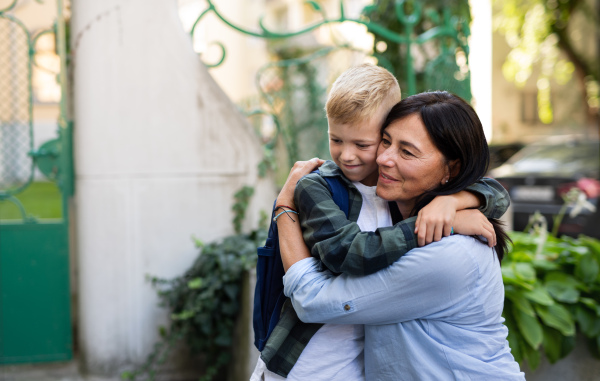 A happy schoolboy hugging his grandmother waiting for him after school outdoors in street.