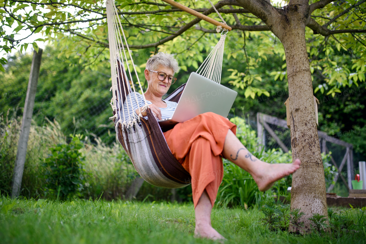 Portrait of active senior woman with laptop working outdoors in garden in hammock, home office concept.