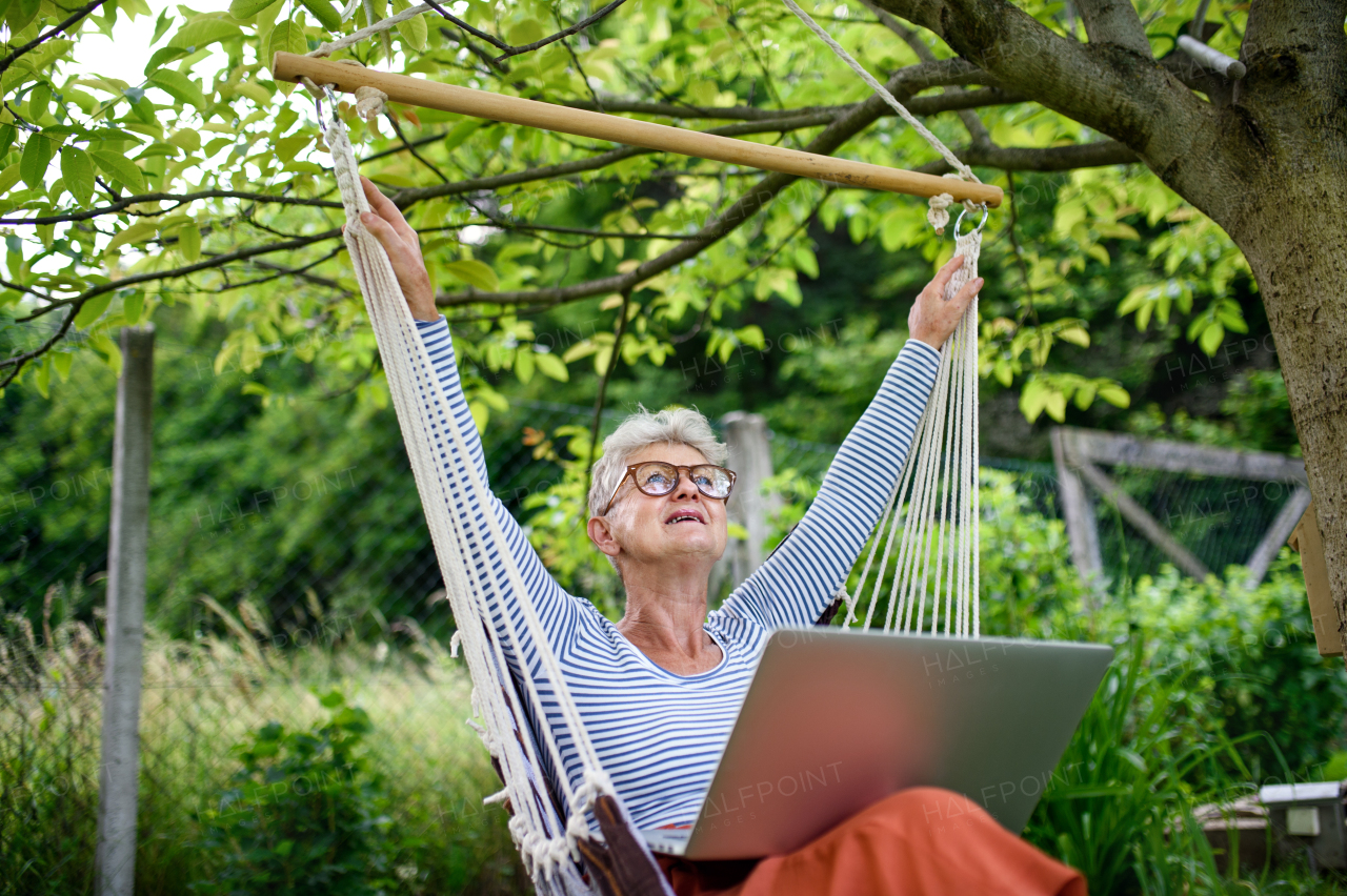 Front view portrait of active senior woman with laptop working outdoors in garden, home office concept.