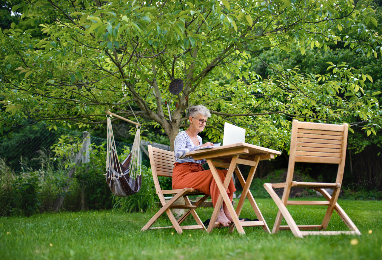 Active senior woman with laptop working at the table outdoors in garden, home office concept.