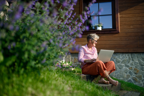 Happy senior woman with laptop working outdoors in garden, home office concept.