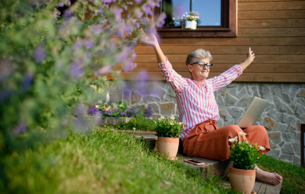 A happy senior woman with laptop working outdoors in garden, home office concept.