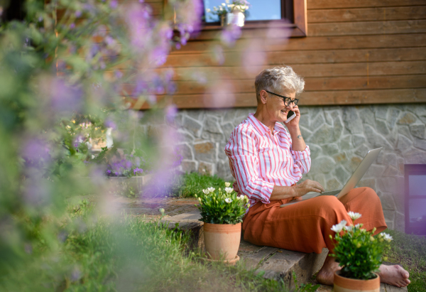 Happy senior woman with laptop, smartphone working outdoors in garden, home office concept.