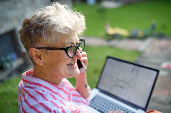 Senior woman architect with laptop and smartphone working outdoors in garden, home office concept.