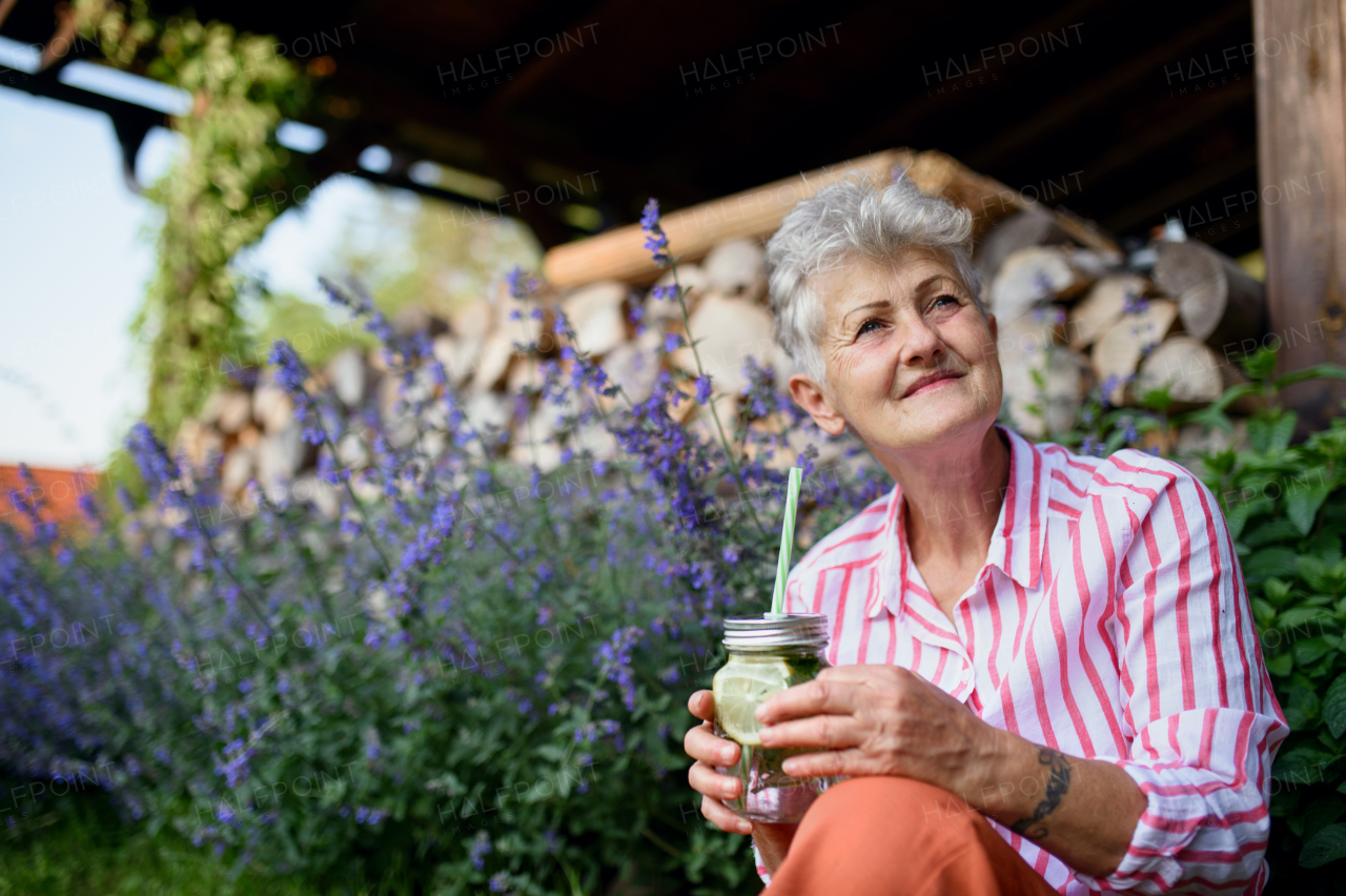 Portrait of senior woman sitting and resting outdoors in garden, holding lemonade drink.