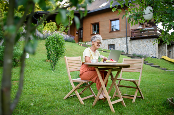 A happy senior woman with laptop working outdoors in garden, home office concept.