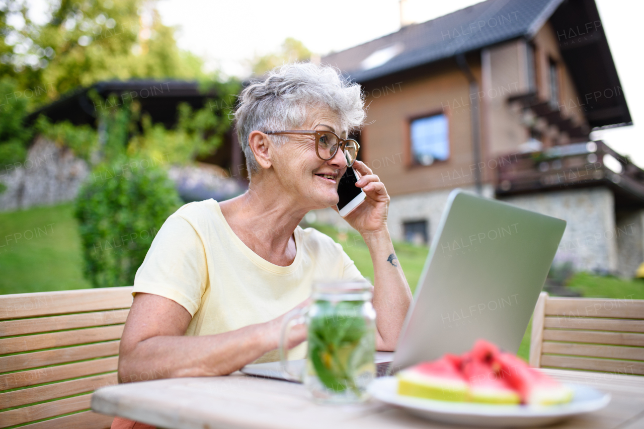 Portrait of happy senior woman with laptop and smartphone working outdoors in garden, home office concept.