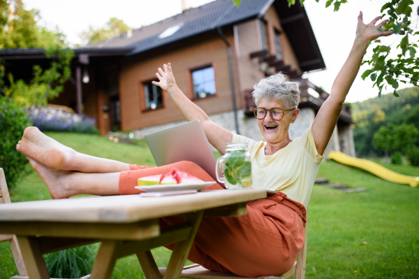 Cheerful senior woman with laptop and feet on desk working outdoors in garden, home office concept.