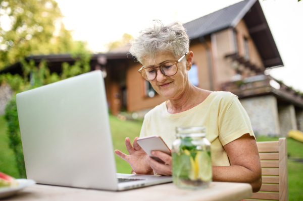 Happy senior woman with laptop, smartphone working outdoors in garden, home office concept.